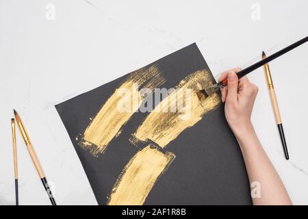 partial view of artist drawing brushstrokes with golden paint in black paper on marble surface Stock Photo