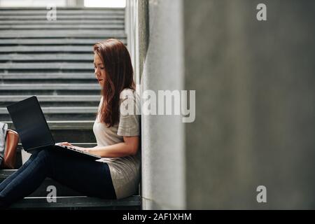 Young Asian woman leaning on wall when resting on steps and reading e-mails on her laptop Stock Photo