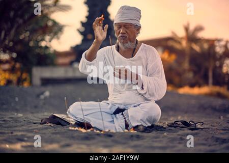 Bali, Indonesia - 12 Dec 2019: Old local balinese man in meditation near the sea. Concept of pray. Adult practice yoga on the Keramas black sand beach Stock Photo