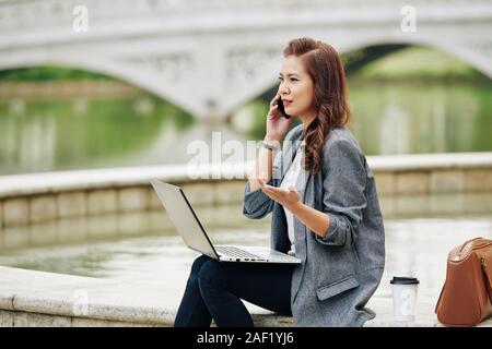 Frowning indignant young businesswoman sitting in park with opened laptop and talking on phone with coworker Stock Photo