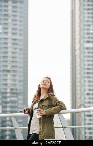 Young Vietnamese woman standing on bridge with smartphone and cup of coffee, closing eyes and enjoying fresh air Stock Photo