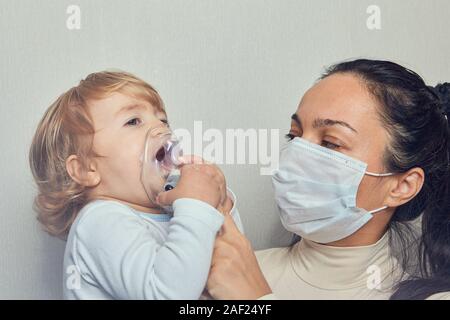White caucasian woman in medical mask is helping her little daughter about 1,5 years old to breathe with special mask, which helps to stop asthma atta Stock Photo