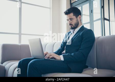 Portrait of his he nice rich imposing attractive focused bearded guy wearing jacket sitting on divan using laptop browsing web surfing in modern Stock Photo