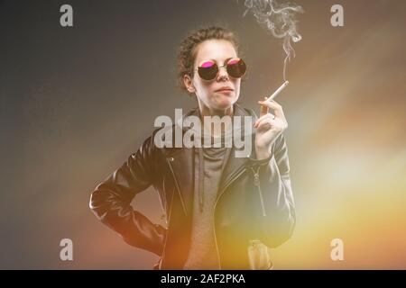 Caucasian girl with rough facial features in a black jacket smokes a cigarette isolated on black background Stock Photo