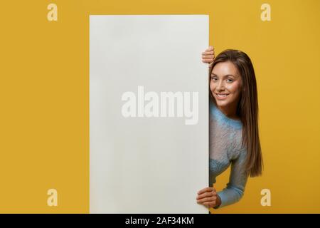 Front view of smiling young pretty girl with long hair presenting white desk on left side. Stylish attractive girl in sweater behind paper sheet posing on yellow studio wall. Concept of advertising. Stock Photo