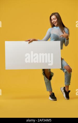 Front view of smiling young stylish brunette girl pointing at white paper sheet in her hand. Fashionable attractive girl with desk posing on yellow studio background. Concept of advertising. Stock Photo