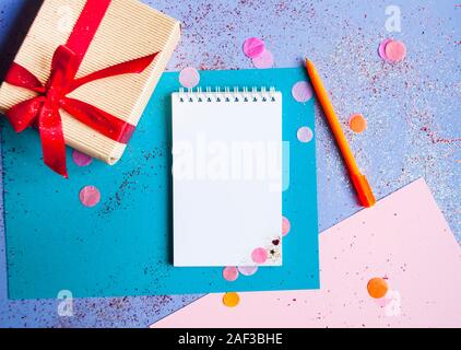 Giftbox bowed with red ribbon on purple background with colorful confetti, giftbox, pen and notebook. New year resolutions concept. Stock Photo