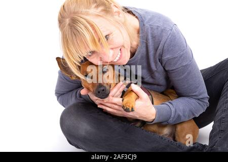 Blonde woman sitting on the floor with her tiny brown dog isolated on white background in the studio.  Small mixed breed of jack russell terrier, germ Stock Photo