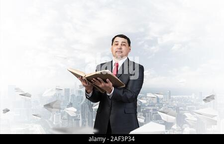 Happy businessman holding open old book. Smiling man in suit standing on downtown background with flying paper planes. Lawyer holding big legal regula Stock Photo