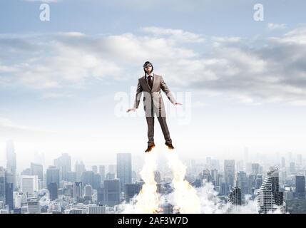 Businessman in suit and aviator hat flying in blue sky as superhero. Corporate manager as superman launching upwards with jet flame above modern city. Stock Photo