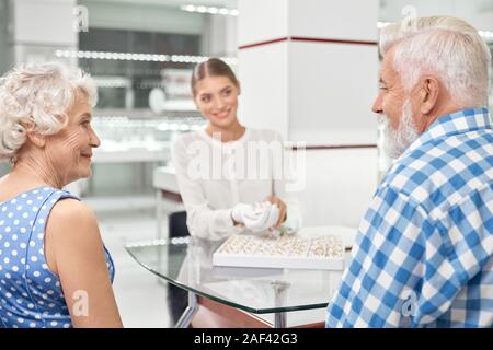 Happy senior romantic couple looking on each other while choosing wedding rings at luxury jewelry market. Professional friendly jeweller helping her customers to buy rings Stock Photo