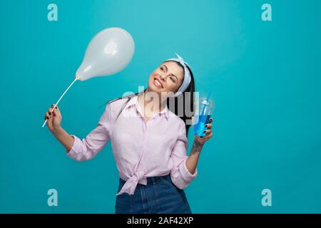 Stylish, happy girl holding plastic cup with blue fizzy drink and white balloon, smiling. Positive model in trendy clothes, stylish hair style posing on blue background. Stock Photo