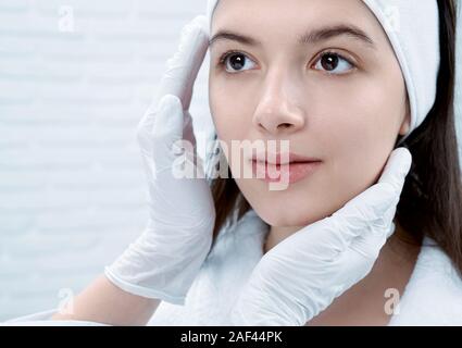 Closeup of charming brunette with pure skin looking at doctor in beauty salon. Hands of female beautician touching and examining skin of client. Concept of treatment and dermatology. Stock Photo