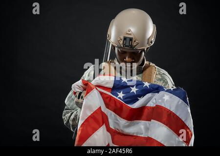 Front view of American soldier wearing uniform and helmet on head holding nationals flag in hands. Ranker sadly looking on American flag. Black isolated background. Stock Photo