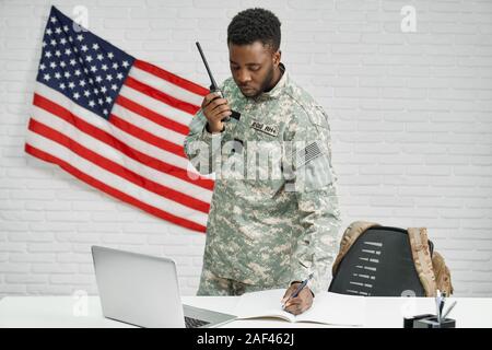 Worker of American army writing documents, using laptop and destination radio remote, talking with colleagues. Young ranker seriously looking and subscribing contract for service in army. Stock Photo