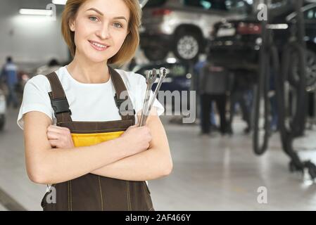 Attractive blonde wearing coverall and white shirt looking at camera and smiling while standing in workshop. Professional female mechanic keeping wrench in hand and posing during working day. Stock Photo