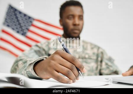 Close up of male soldier hand holding pen and writing document. American ranker seriously looking and subscribing contract for service in army. American flag on background. Stock Photo