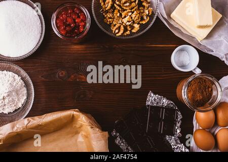 Overhead view of ingredients for homemade baking chocolate brownie cake with walnuts and dried cherries on dark wooden table, flat lay. Stock Photo