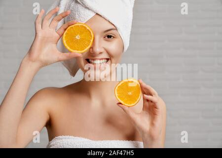 Front view of cheerful woman wearing towel after shower keeping fresh oranges and looking at camera. Attractive woman doing rejuvenating masks and caring about face and body skin. Concept of beauty. Stock Photo