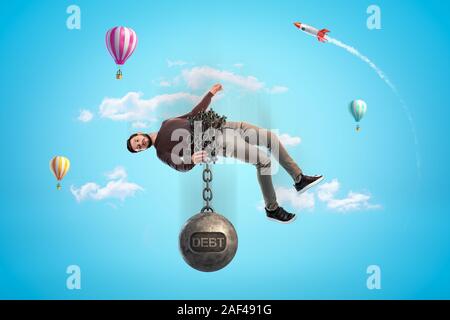Man in blue sky bound with chains to big metal ball that reads 'DEBT' and is pulling him down. Having financial problems. Heavy debts. Going bankrupt. Stock Photo