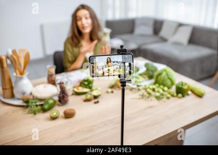 Young woman recording on a smart phone her vlog about healthy eating. Sitting at the table with lots of green vegan food ingredients at home Stock Photo