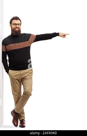 Full length portrait of a bearded man in a turtleneck sweater leaning on a wall and pointing to the side isolated on white background Stock Photo