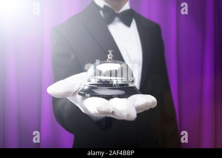 Midsection Of Male Waiter Holding Service Bell On Hand Against Purple Curtain Stock Photo