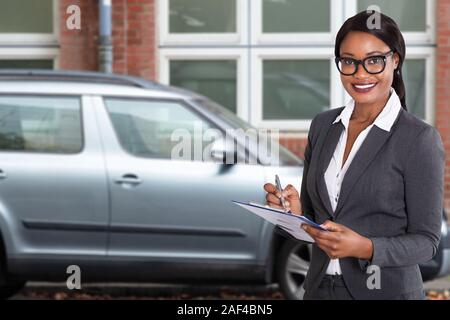 African Woman Holding Papers On Clipboard With Pen Standing Near Parked Car Outside Apartment Stock Photo