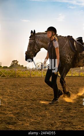 Beautiful girl jockey stand next to her horse wearing special uniform on a sky and green field background on a sunset Stock Photo