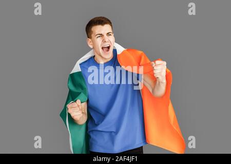Freestyle. Young man standing isolated on grey with irish flag supporting team shouting excited Stock Photo