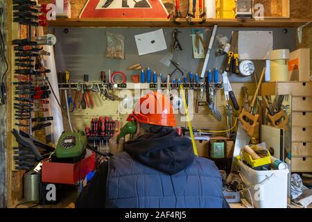 Craftsman with a construction helmet on the phone with customers, sitting in front of a workbench overfilled with rubbish Stock Photo