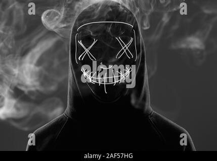 Close-up portrait of an anonymous man hiding his face behind a scary neon mask in a colored smoke. Studio shot Stock Photo