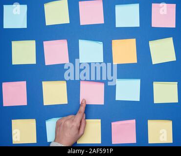 female hand and a lot of empty paper multi-colored square stickers on a blue background, concept of choosing a strategy with the specified finger Stock Photo
