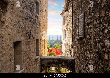 The red umbrellas of a cafe patio and the French Alpes-Maritimes countryside are visible from an alleyway in medieval Saint Paul de Vence, France Stock Photo