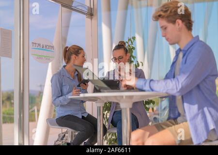 Two women talk to each other while using phones and a tablet, with an out of focus man at the front Stock Photo