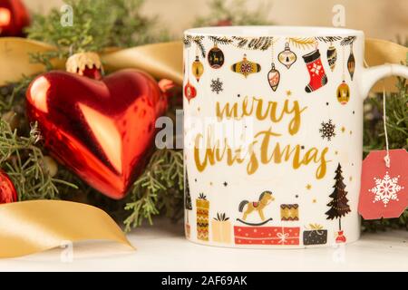 Close up of a Christmas mug with hanging empty tea label with around pine branches, red baubles and satin gold ribbon with bokeh effect Stock Photo