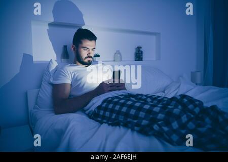 Portrait of his he nice attractive focused guy lying on bed using cell sending sms browsing web site at night late evening home dark illuminated room Stock Photo