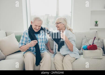 Portrait of his he her she two nice friendly lovely cheerful cheery people sitting on divan granny wrapping granddad in new scarf in light white Stock Photo