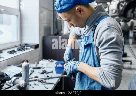 Repairman in car service wearing blue uniform and protective rubber gloves working near table with a lot of tools on it,preparing details to replace t Stock Photo