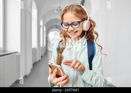 Beautiful little school girl with blonde curls wearing eyeglasses and blue blouse using smartphone and smiling. Pretty girl on corridor, holding call Stock Photo