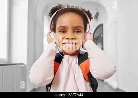 Front view of positive and smiling little African girl wearing big pink headphones looking at camera. School girl standing on corridor and listening m Stock Photo