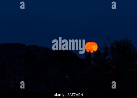A beautiful full red moon rises from behind a hill with trees Stock Photo