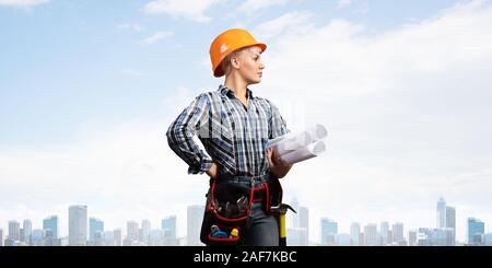 Attractive female engineer in hardhat standing with technical blueprints. Portrait of young architect in checkered blue shirt on background of citysca Stock Photo