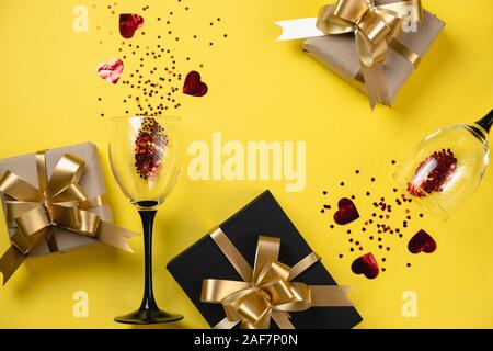 Valentine's Day background. Two clinking wine glasses, various handmade gift boxes decorated with red heart confetti on yellow background. Valentines Stock Photo