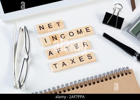 Net income after taxes concept with letters on cubes. Still life of office workplace with supplies. Flat lay white surface with tablet computer and ca Stock Photo