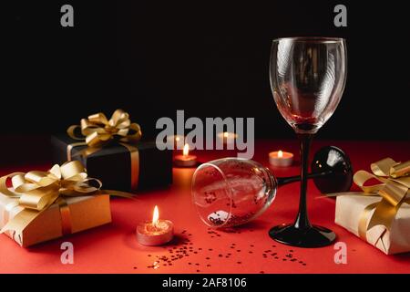 Valentine's Day background. Two clinking wine glasses, various handmade gift boxes decorated with confetti, candle on red and black background. Valent Stock Photo