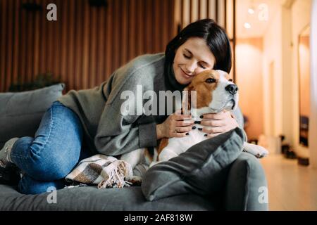 A woman lies on a sofa next to a beagle dog and hugs him.