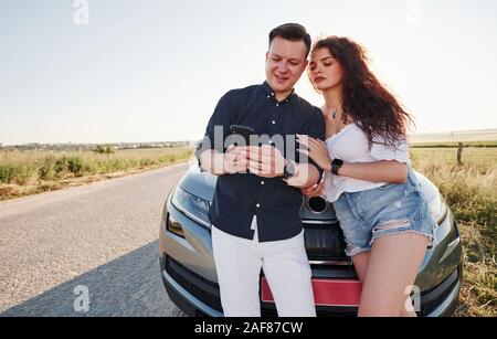 Using smartphone. Lovely couple near their new modern car at weekend time Stock Photo