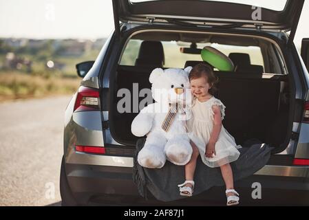 Cute child with white teddy bear toy sits on the back of automobile Stock Photo