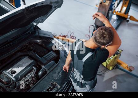 Sweaty and tired. Man in grey uniform repairs white automobile indoors Stock Photo